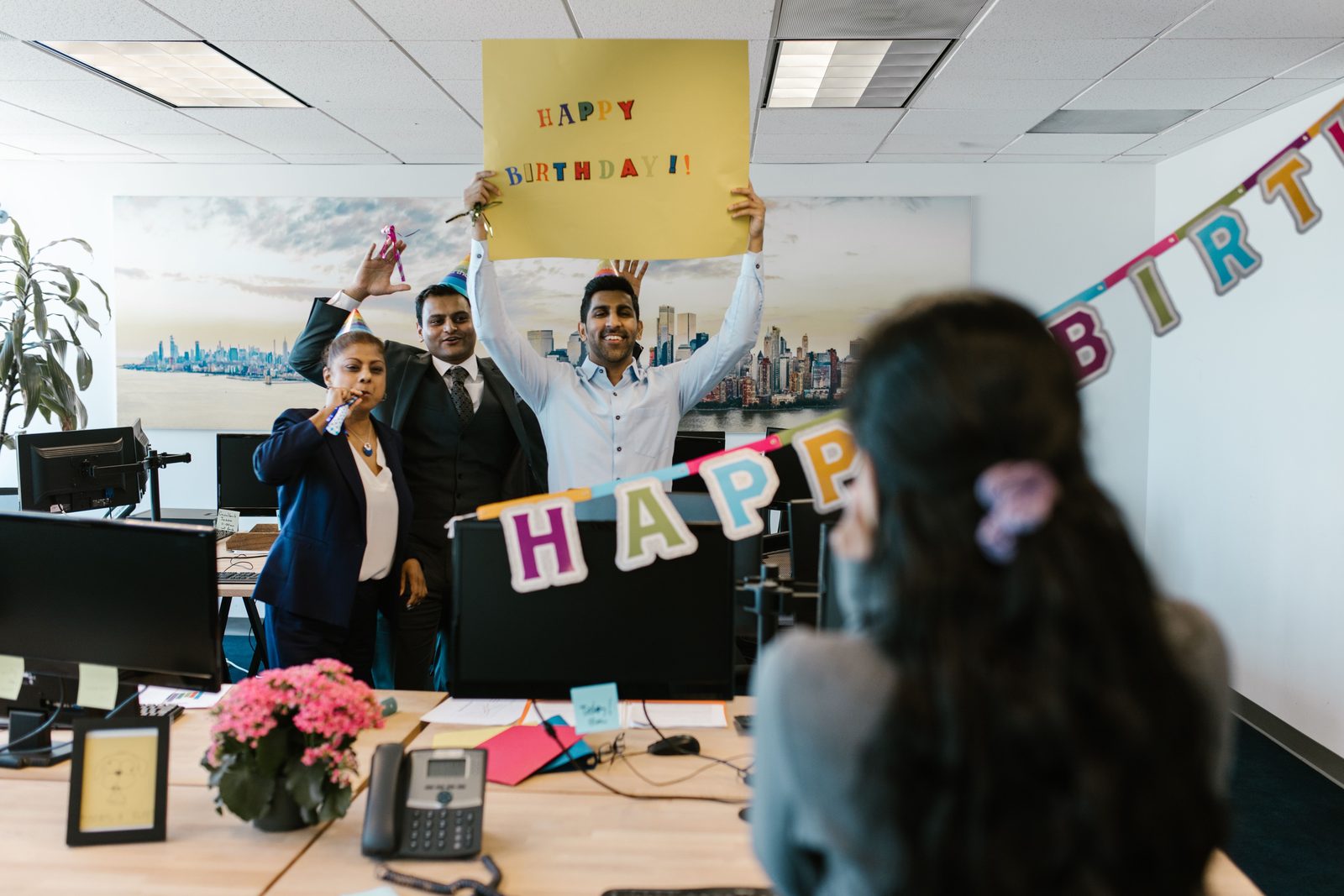Coworkers in party hats surprising a colleague with a Happy Birthday poster at her desk